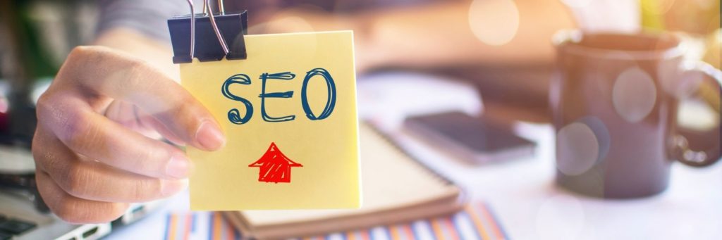 Person holding a sticky note with "seo" written on it and an arrow pointing upwards, symbolizing search engine optimization improvement as part of a website strategy.