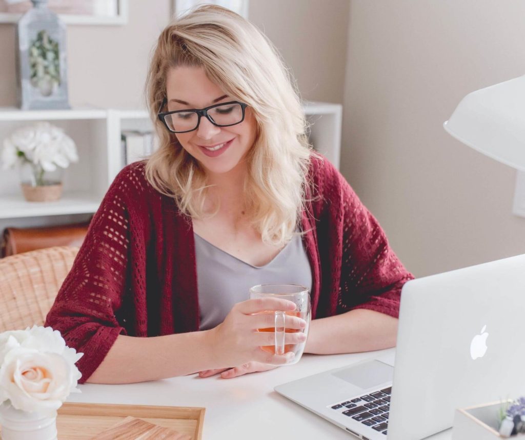 A woman wearing glasses, holding a glass of water, and smiling while looking at her laptop in a well-lit room as she reviews her website strategy with elliot olson from portland, oregon.