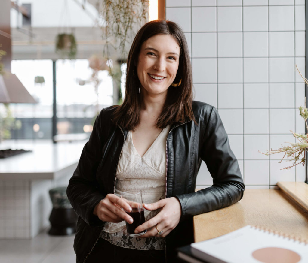 A smiling woman sitting at a table in portland, oregon, wearing a black leather jacket, holding a glass.