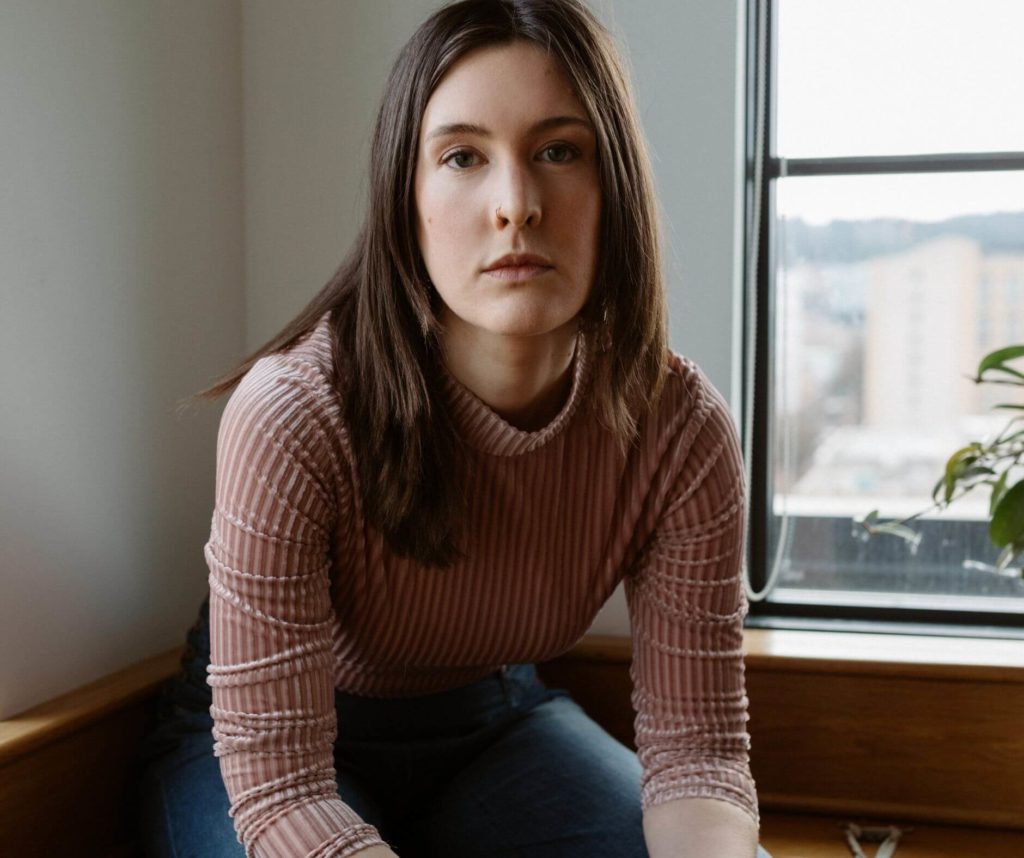 A woman in a ribbed top sitting by the window, looking thoughtfully at the camera for studio anansi's website strategy.