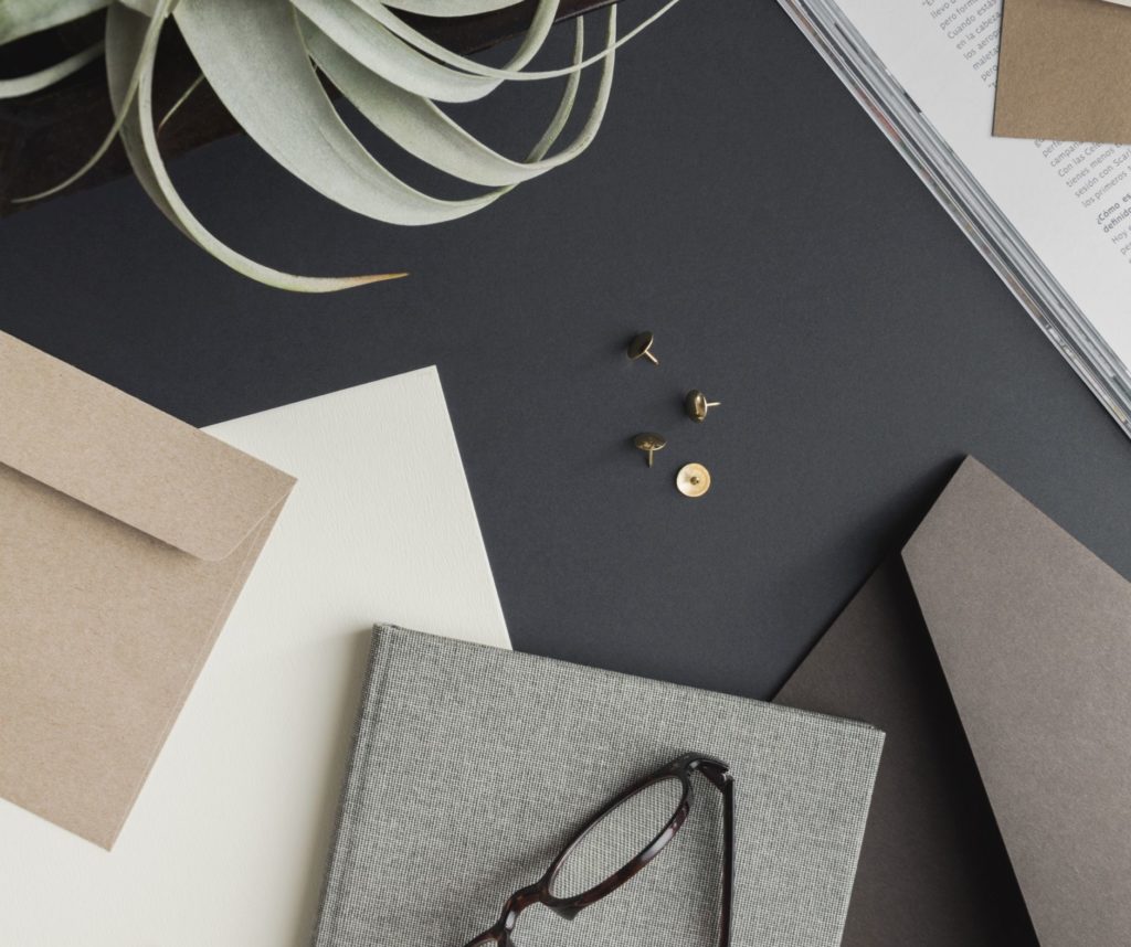 Flat lay of studio anansi stationery items including envelopes, an open book, sprigs of a plant, pushpins, and eyeglasses on a neutral-colored surface.