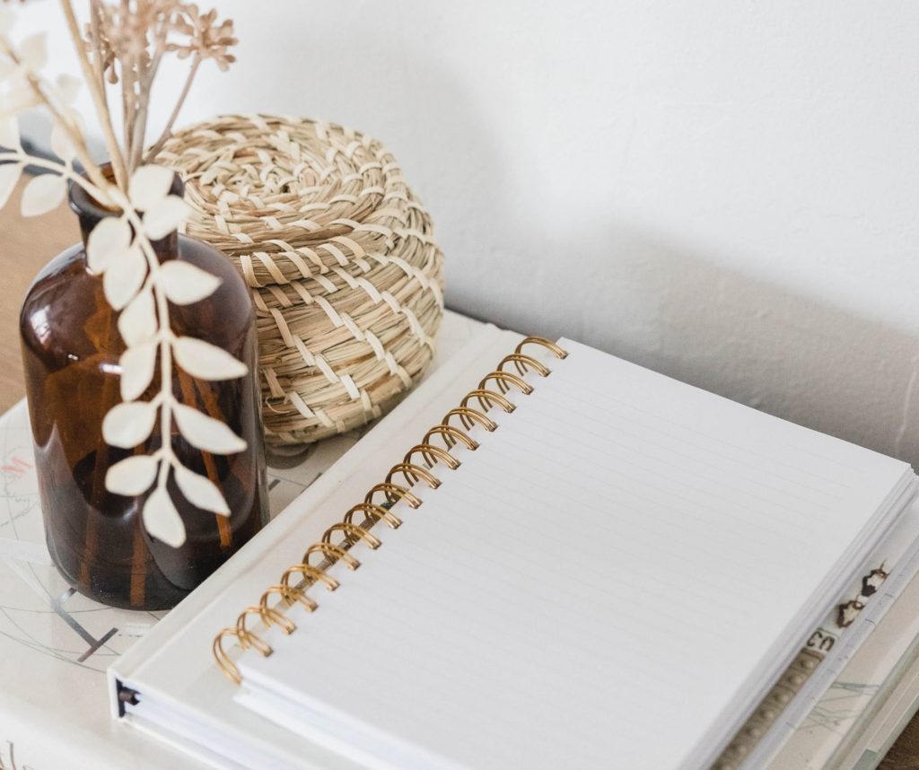 An open blank notebook with a spiral binding on a white surface, next to a woven basket and a brown vase with dried plants, photographed by elliot olson at studio anansi in portland, oregon.