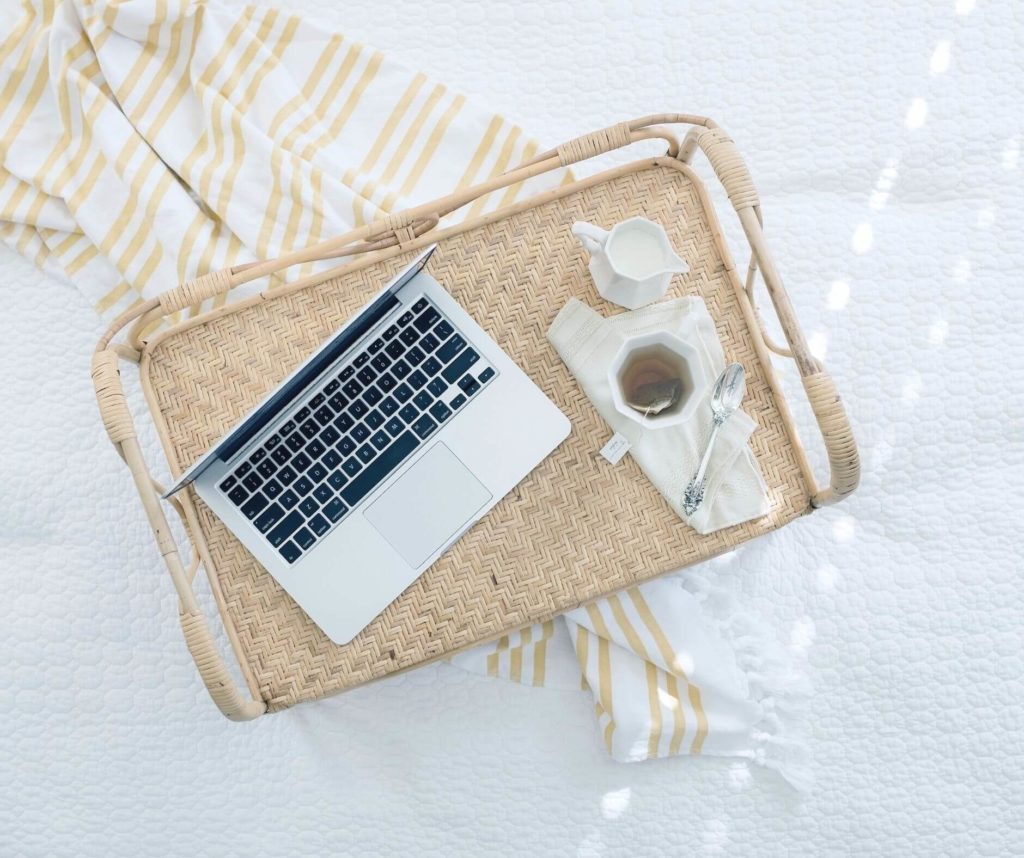 A laptop on a wicker tray beside a cup of coffee and a napkin on a bed with white linens and yellow stripes, curated by studio anansi.