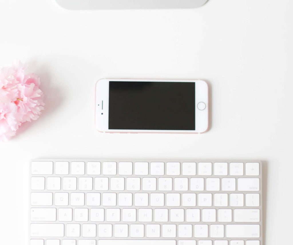 White desk with a smartphone lying next to a keyboard, a pink flower, and materials for website design.