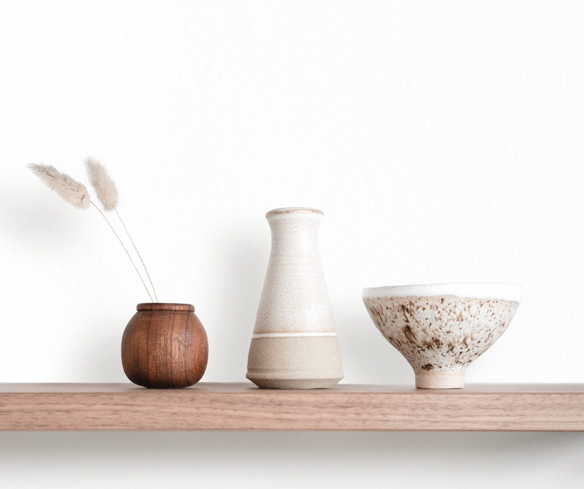 Three ceramic vessels by Studio Anansi on a wooden shelf against a white wall, with dried grasses in the smallest one.