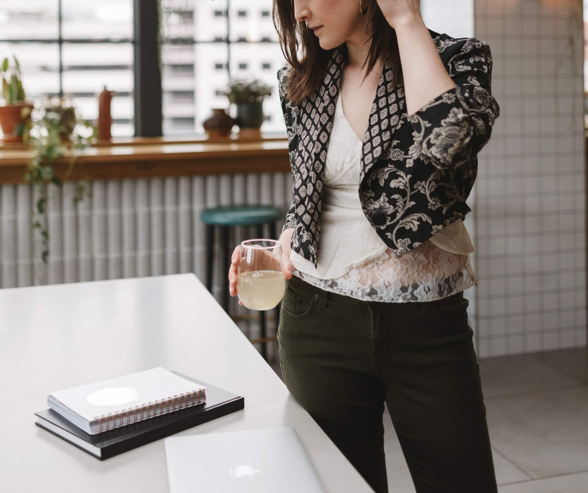 A woman in a patterned blazer and green trousers stands by a table holding a glass, with a laptop and notebook nearby, working on website design.