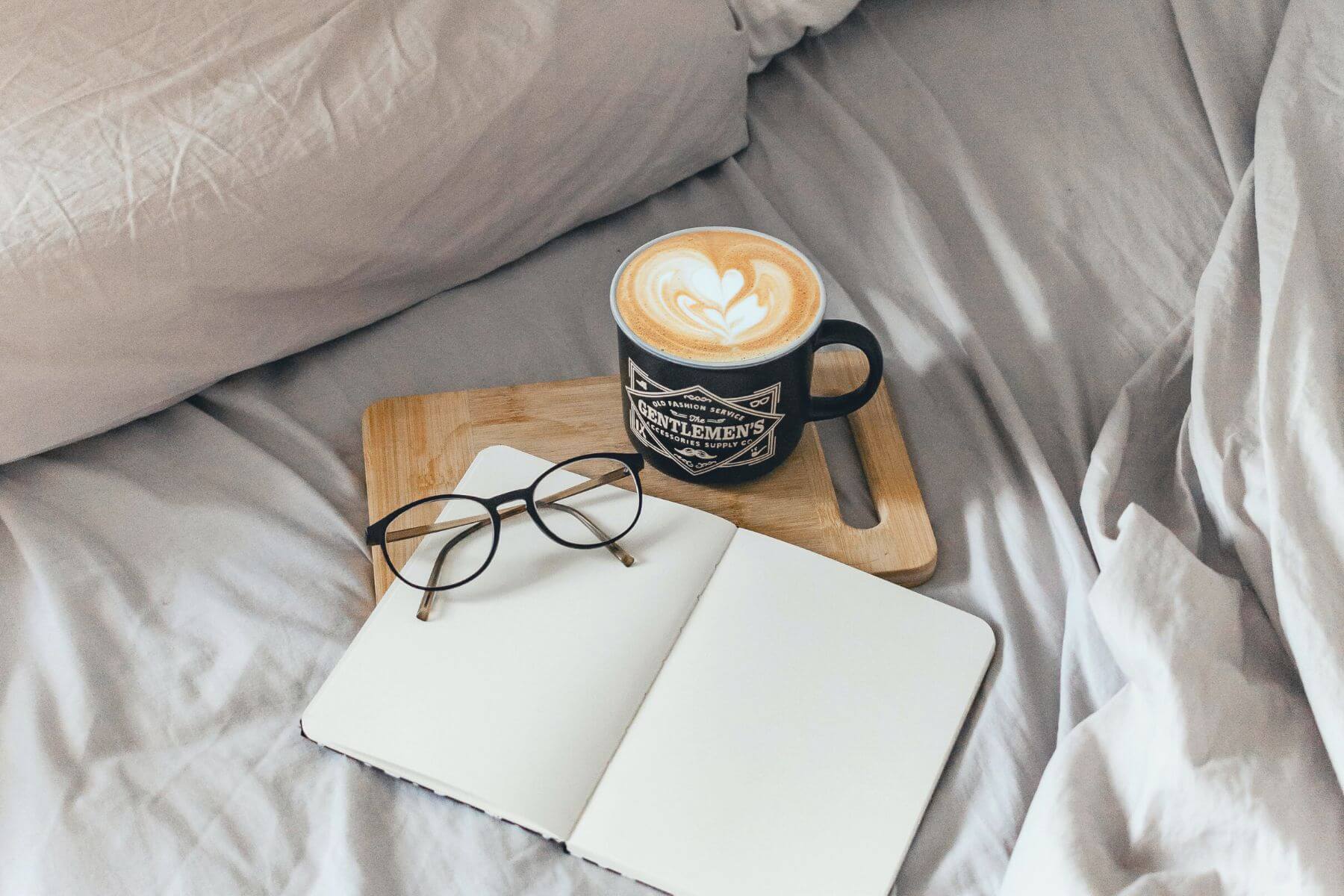 A cup of coffee and a notebook are resting on a bed.